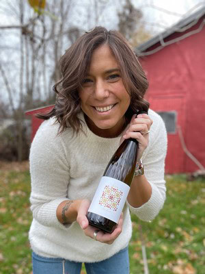 Woman holding a bottle of Bos Wine outdoors, smiling, with a red barn in the background.