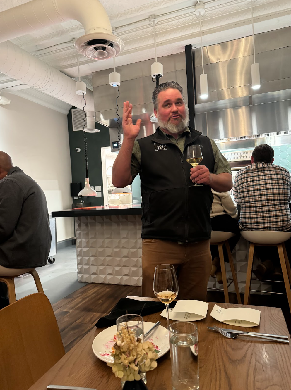 Man with gray beard holding a glass of white wine, speaking to guests in a modern winery setting.
